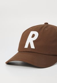Brown baseball cap featuring a prominent white embroidered 'R' on the front, with a curved brim and textured fabric surface.