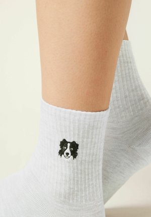 Ankle-high white socks with ribbed texture featuring a small black and white dog face embroidered near the ankle on a person's legs.