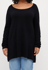 Kroezigharig persoon in een lange zwarte trui met een ronde hals en lichtblauwe jeans, staande tegen een effen witte achtergrond.