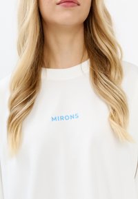 Witte sweatshirt met blauwe geborduurde tekst "MIRONS." De stof lijkt zacht, met een klassiek ronde halsontwerp.