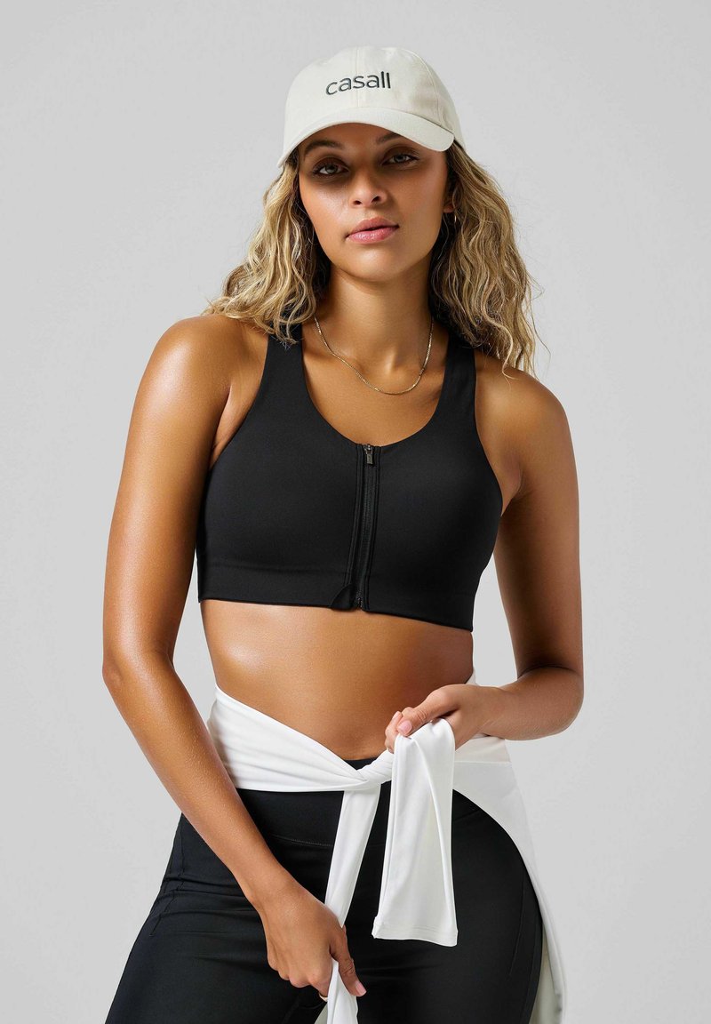 Czarny sportowy stanik na zamek z szerokimi ramiączkami i gładką teksturą, zestawiony z białym materiałem związanym w talii. Swobodne beżowe czapka z logo.