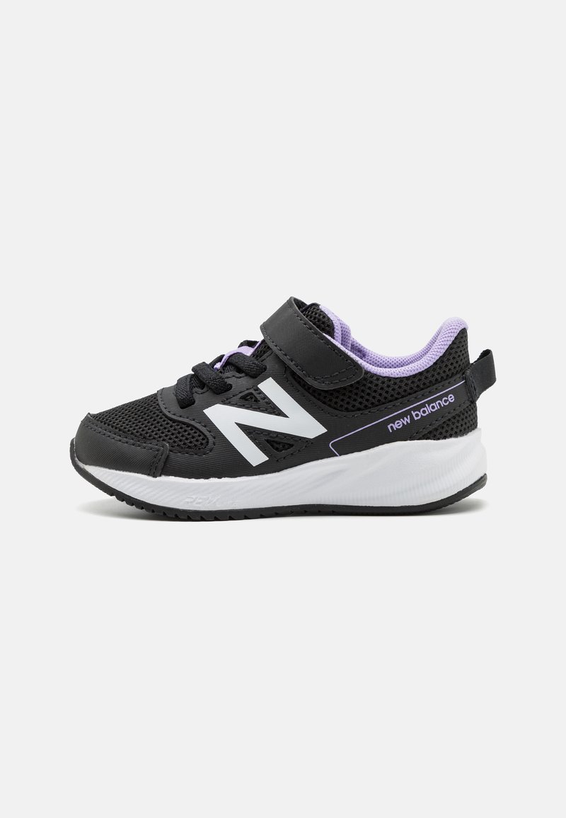 Zapatilla deportiva negra con parte superior de malla, correa ajustable, acentos morados y suela blanca. Presenta un prominente logo blanco en el lateral.