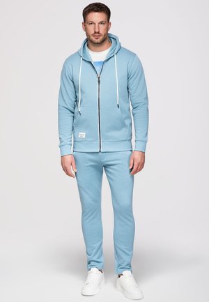 Homme debout portant un sweat à capuche zippé bleu clair, un pantalon de jogging assorti, des baskets blanches et un t-shirt blanc avec un motif bleu.