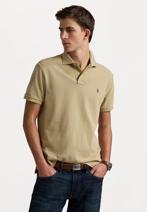 Polo beige avec une finition texturée, patte de boutonnage à deux boutons, et un petit logo bleu sur la poitrine, associé à un jean bleu foncé et une ceinture marron.