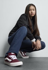 Červené plátené skateboardové topánky s bielymi akcentmi a šnúrkami; štruktúrovaná gumená podrážka. Modré rifľové džiny a čierna bomber bunda. Sivé pozadie.