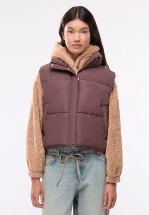 Donna che indossa un gilet imbottito marrone sopra una felpa beige pelosa con cappuccio, abbinata a jeans azzurri legati in vita con un laccio.
