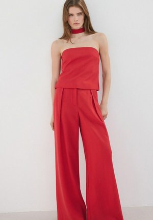 Femme debout contre un mur blanc, portant un haut rouge sans bretelles, un pantalon large assorti et un collier rouge, avec une expression neutre.