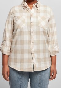 Persona che indossa una camicia a quadri beige e bianca con maniche arrotolate e jeans di denim azzurro chiaro su uno sfondo neutro.