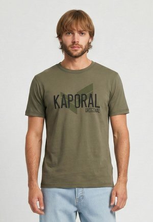 Homme aux cheveux châtain clair et à la barbe, portant un t-shirt Kaporal Original vert olive et un jean bleu clair, debout devant un fond uni.