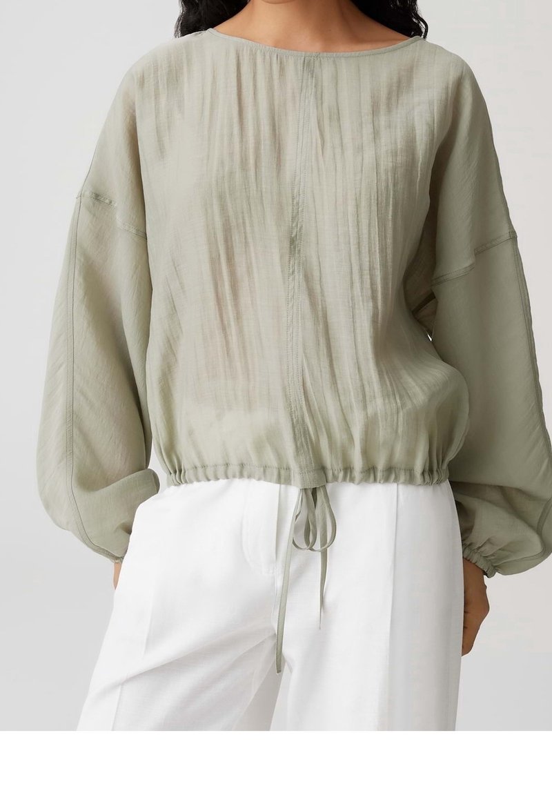 Blouse vert clair à manches longues avec ourlet froncé et lien ajustable, associée à un pantalon blanc taille haute à jambes larges.