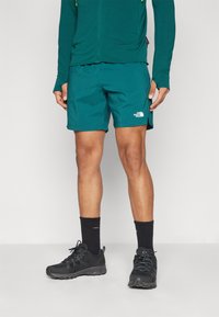 Shorts de course teal avec un logo blanc, associés à un haut long-sleeve teal et des chaussures de trail noires, présentent un tissu texturé et un design ventilé.