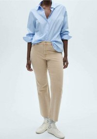 Personne portant une chemise à boutons bleu clair avec manches retroussées, pantalon beige à jambes droites, chaussettes blanches et baskets blanches, debout sur un sol gris.