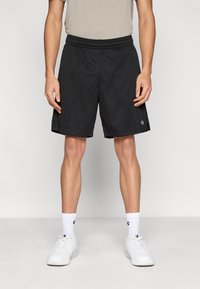 Μαύρα αθλητικά σορτς από δίχτυ με ελαστική μέση. Διαθέτουν ένα μικρό λογότυπο στη κάτω αριστερή πλευρά. Φοριούνται με λευκά αθλητικά παπούτσια και κάλτσες.