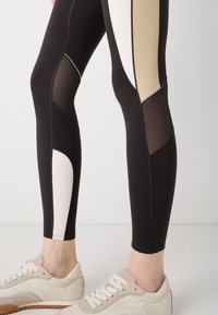 Leggings neri con pannelli beige e bianchi, caratterizzati da inserti in rete e cuciture a contrasto. Abbinati a sneakers chiare.