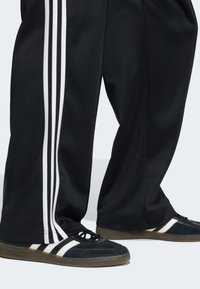 Crne sportske hlače s bijelim prugama u tri linije. Nošene s crnim cipelama od brušene kože koje imaju bijele i crne pruge te gumenim potplatom.