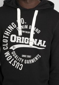 Felpa nera con cappuccio e stampa grafica circolare bianca. Caratterizzata da coulisse, polsini a coste e texture morbida. Il marchio include le scritte "Original" e "Custom Garments."