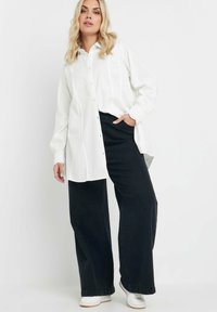 Camicia bianca button-up in tessuto testurizzato, maniche lunghe e vestibilità rilassata, abbinata a pantaloni neri a vita alta con gamba larga e sneakers bianche.