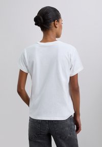 Kobieta z ciemnymi włosami upiętymi w kok, ubrana w gładki biały t-shirt z krótkim rękawem i wyblakłe czarne dżinsy, odwrócona tyłem w stosunku do jednolitego jasnego tła.