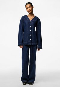 Denim odijelo u tamnoplavoj boji, s V-izrezom, velikim dugmadi i dugim rukavima. Hlače slobodnog kroja s raširenim oblikom.
