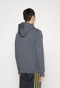Sudadera gris con capucha y mangas largas, con un corte relajado y detalles de costura visibles. Llevada con pantalones negros que tienen rayas verticales verdes.