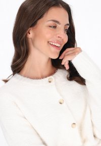 Cardigan alb tricotat cu guler rotund, model texturat și trei nasturi crem. Modelul își atinge manșeta, subliniind materialul moale.