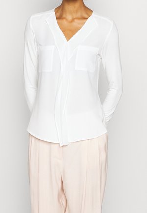 Mujer con blusa blanca de manga larga y escote en V, con dos bolsillos en el pecho y pantalones plisados de color beige claro.