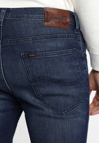 Tummansinisiä farkkuja, joissa on sileä pinta, ja joissa on ruskea nahkainen takapatch, jossa on kohokuvioituna "Lee" sekä erottuva takataskustitching.