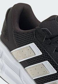 Czarny but sportowy z siateczkową cholewką i zamszowymi akcentami, z wyraźnymi białymi paskami na wierzchu oraz teksturowanymi detalami w okolicy sznurowadeł.