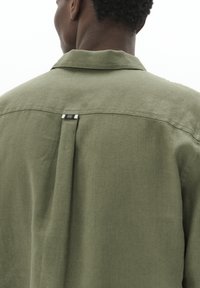 Camicia verde oliva con colletto, caratterizzata da una piega trasversale sul retro e un piccolo etichetta a righe vicino alla scollatura. Realizzata in tessuto testurizzato.