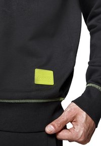 Czarna bluza z miękkiego materiału z zielonymi przeszyciami i kwadratową żółtą metką z logo "Aramco Formula One Team."