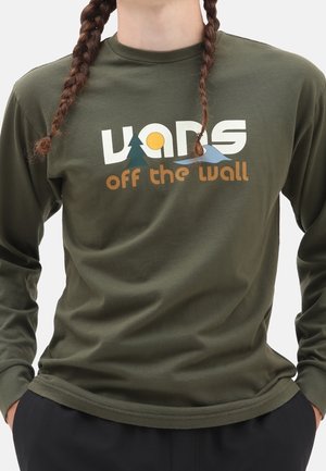 Osoba z warkoczykami, ubraną w oliwkową koszulę z długim rękawem, na której znajduje się stylizowane logo Vans z grafiką drzewa, słońca i fali.