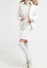Vit puffjacka med en pälsfodrad huva, stickad tröja, vit kjol, strumpor till knäna och vita sneakers med perforerade detaljer.