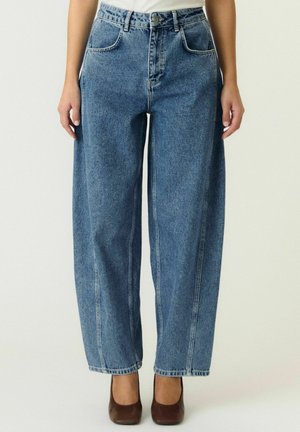 Personne portant un jean bleu taille haute à jambes larges, une chemise blanche et des chaussures à talons marron, debout devant un fond uni clair.