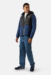 Granatowo-czarna kurtka zapinana na zamek z kapturem, pikowana górna część, gładka dolna część oraz biały dziergany sweter pod spodem. Nosi się ją z jeansami typu cargo.