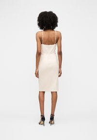 Femme aux cheveux bouclés portant une robe beige sans manches jusqu'aux genoux et des sandales noires à talons hauts, debout, dos à la caméra.