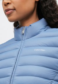 Γυναίκα που φοράει ένα ελαφρύ μπλε ζακέτα με γέμισμα και όρθιο γιακά, με το λογότυπο "Calvin Klein" στο στήθος, με το φερμουάρ πλήρως κλειστό.