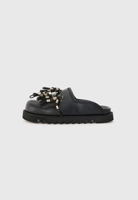 Pantofii negri tip slip-on, cu o talpă din cauciuc texturat, având accente metalice și bucle pe partea superioară pentru detalii decorative.