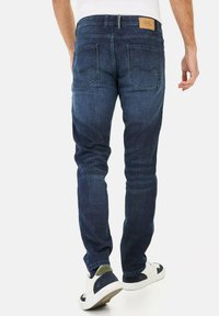 Jean en denim bleu foncé avec une coupe slim, cinq poches, coutures visibles et une étiquette en cuir sur la taille. Porté avec des baskets blanches.