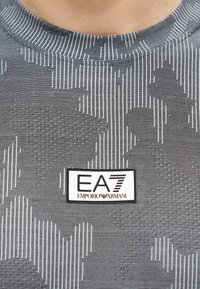 Κοντινό πλάνο ενός γκρι πουκάμισου με σχέδιο και λευκή ορθογώνια ετικέτα στο κέντρο εμπρός, με το λογότυπο «EA7 Emporio Armani».