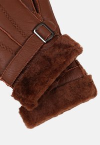 Guanti in pelle marrone con un morbido polsino in faux fur marrone, dotati di una fibbia metallica rettangolare e cuciture dettagliate sulla pelle.