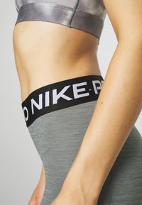 I leggings sportivi Nike Pro grigi presentano una ampia vita nera con scritte bianche. Il tessuto ha una leggera texture ed è aderente.
