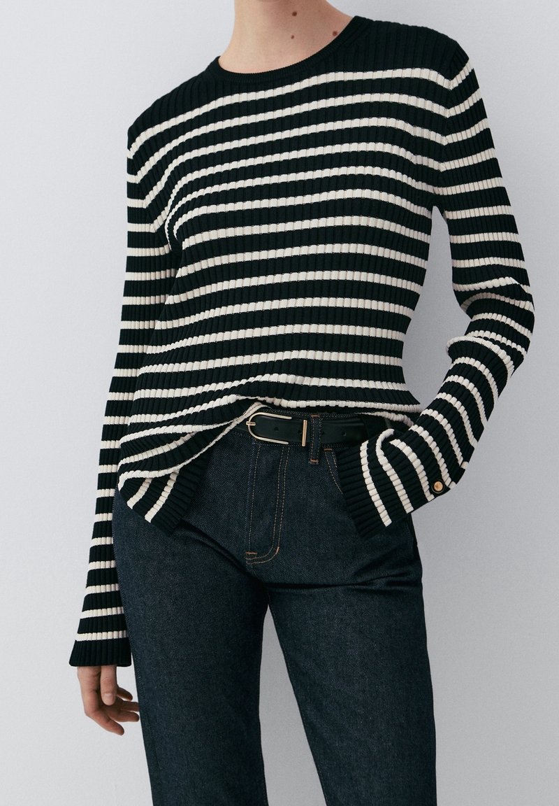 Personne portant un pull côtelé à rayures horizontales noires et blanches, rentré dans un jean foncé taille haute avec une ceinture noire.