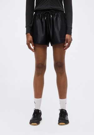 Persona che indossa pantaloncini sportivi neri, maglia nera a maniche lunghe, calzini bianchi e scarpe da ginnastica nere, in piedi davanti a uno sfondo semplice.