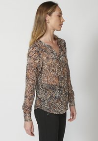 Blusa trasparente a manica lunga con un motivo di foglie multicolore in marrone, crema e blu navy. Presenta una chiusura con bottoni e un orlo arrotondato.