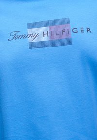 Μπλε ύφασμα με το λογότυπο Tommy Hilfiger που διαθέτει ορθογώνιο με σχέδιο πλέγματος σε ναυτικό μπλε, λευκό και κόκκινο δίπλα σε στυλιζαρισμένο κείμενο.