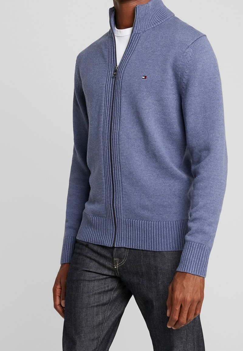 Blå stickad jacka med hög krage, ribbade muddar och fåll, med en liten logotyp på bröstet och en strukturerad tyg.