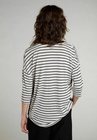 Maglietta a righe, ampia e comoda, in crema e nero, con maniche a tre quarti e orlo curvo. Accenti decorativi sulle spalle.