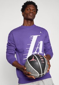Sweatshirt roxo com gráfico branco "LA" e pequeno logotipo; calças de fato cinzas; segurando uma bola de basquete preta texturizada com acentos vermelhos.