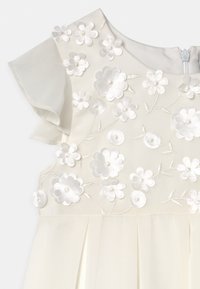 Vit klänning med broderade blommor i vitt och silver, med plisserad kjol och genomskinliga, svajande ärmar. Dragkedja i ryggen.