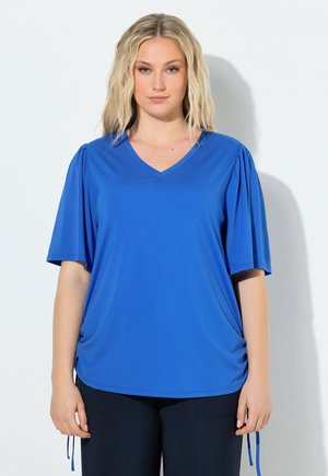 Blonde vrouw in een koningsblauw V-hals top met korte pofmouwen, staand tegen een effen lichte achtergrond.
